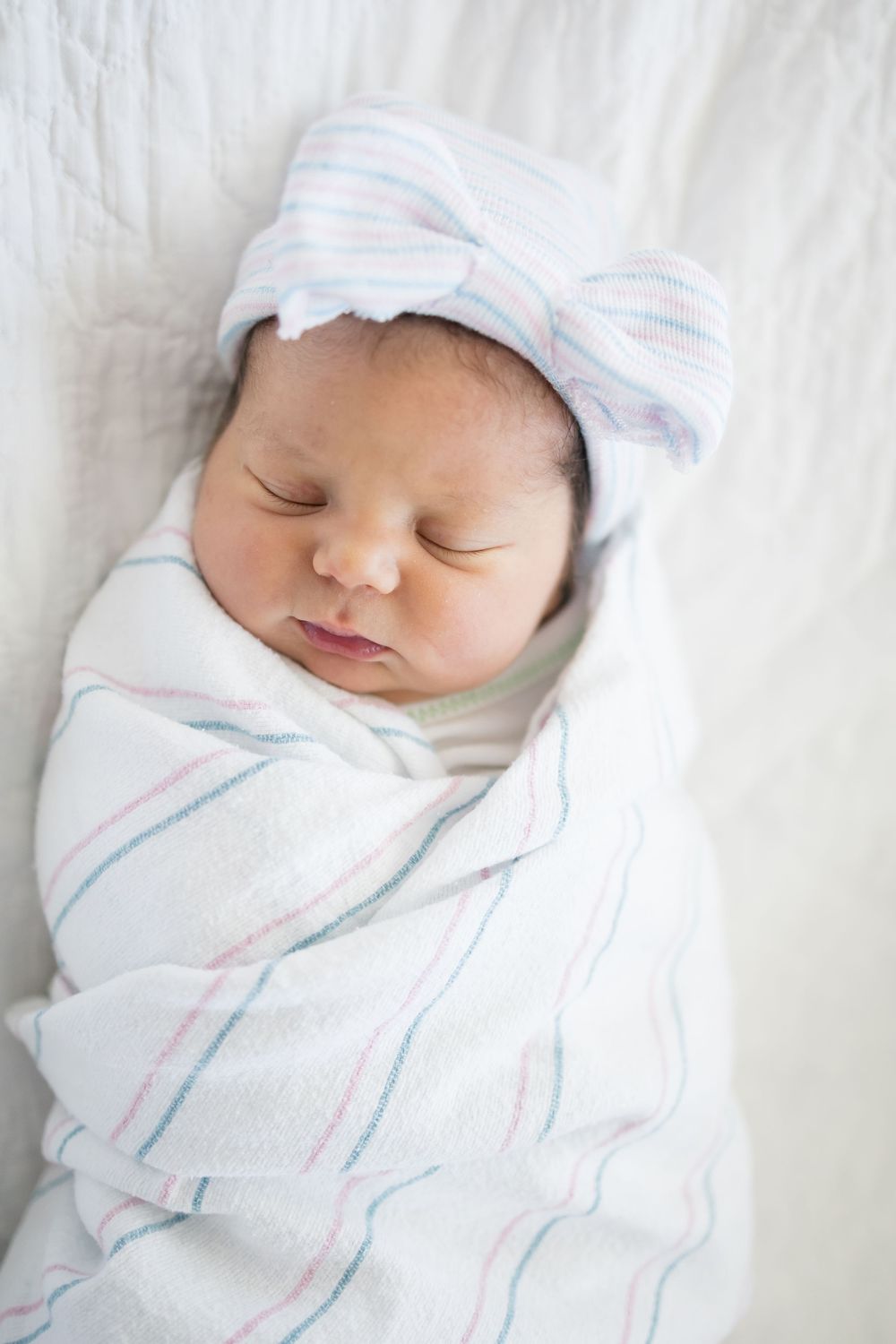 Peaceful newborn baby swaddled in white striped blanket with bow hat sleeping during her Fresh 48 session at Fort Sanders Medical Center.