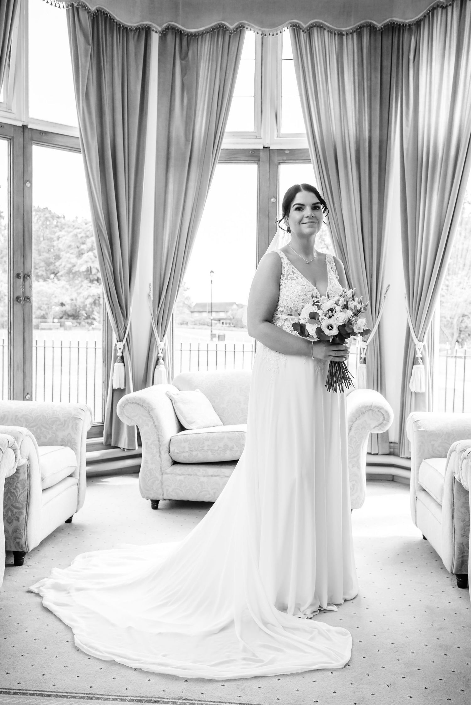 Elegant bride in white gown holding bouquet stands before large curtained windows in a classic interior.