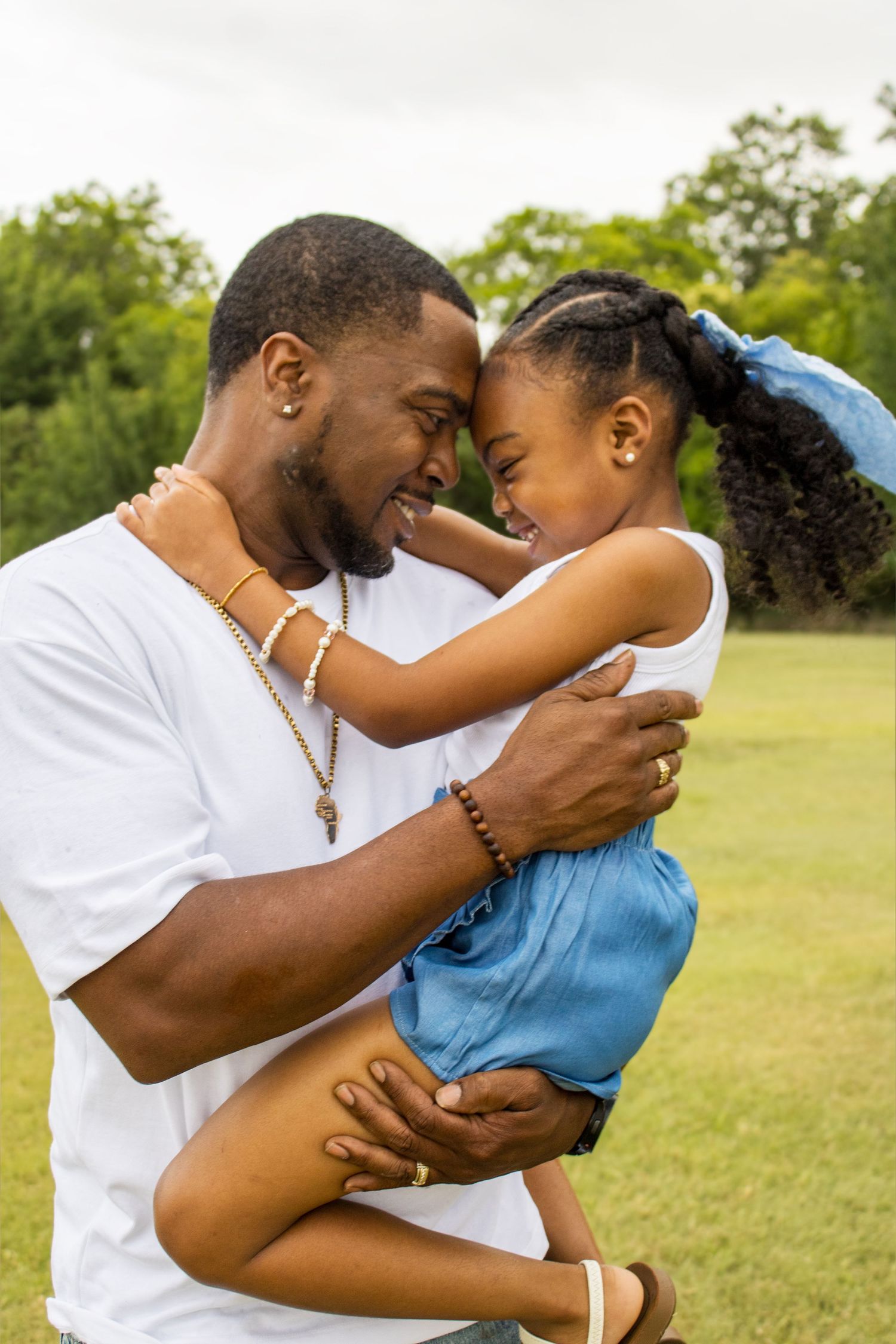 A smiling Black father holds his laughing young daughter close in a green outdoor park.