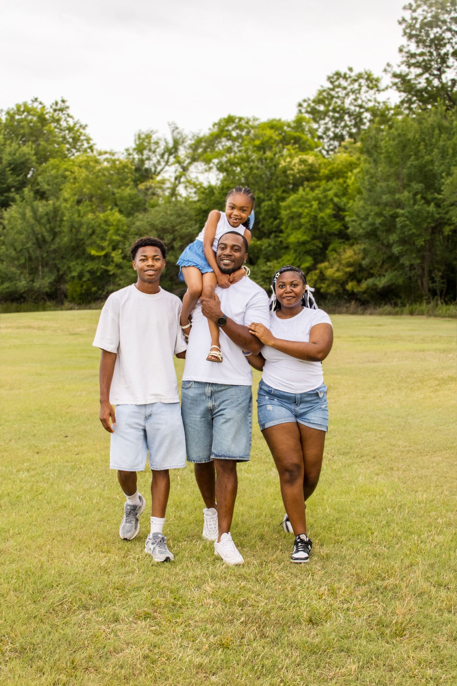 A happy Black family of four poses together on a green lawn with trees in the background.