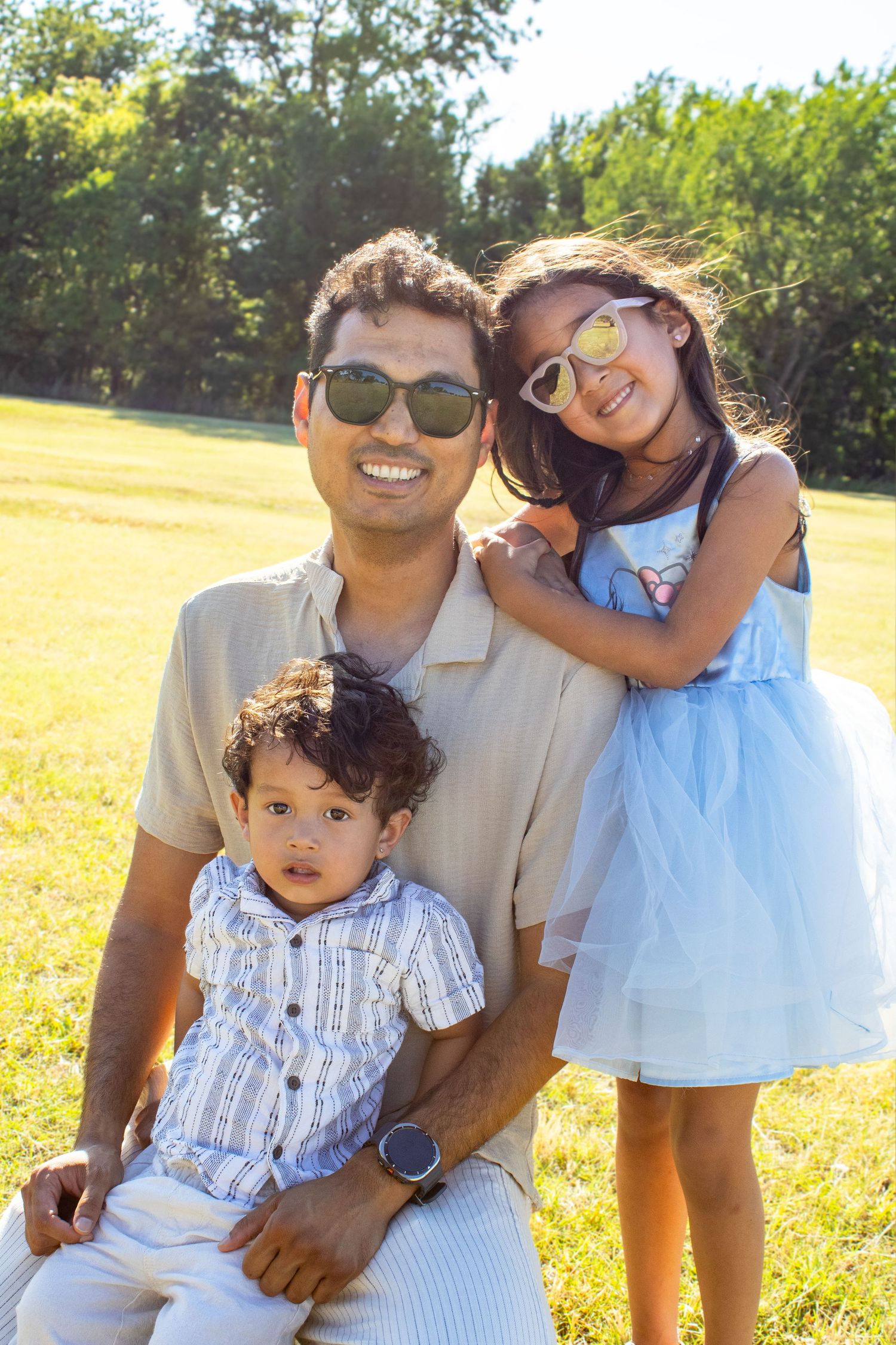 A smiling father with sunglasses sits with his two young children in a sunny green park.