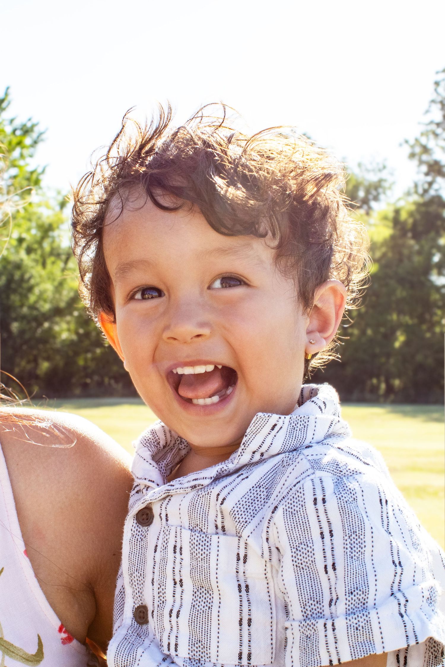 A cheerful toddler boy in a striped shirt laughs brightly in a sunny outdoor setting.