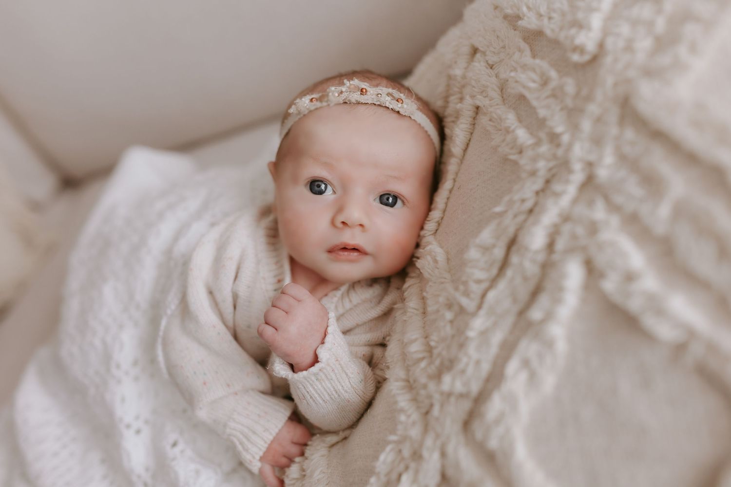 Adorable newborn baby girl in cream knit outfit and floral headband nestled in soft white blanket.