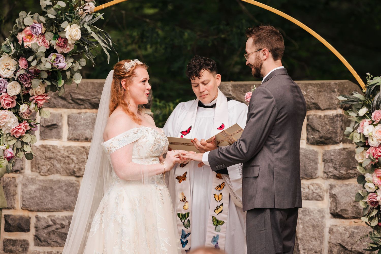 Bride and groom exchanging rings beneath the floral arch, officiant reading the vows aloud as the couple smiles at each other.