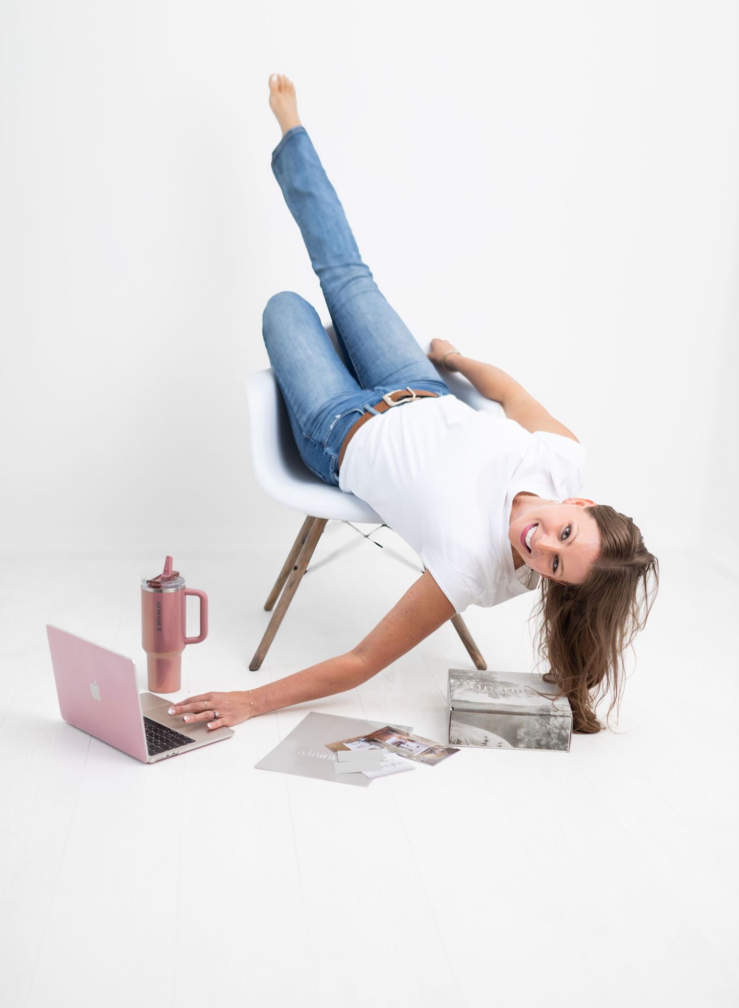 Woman in jeans and white tee balances sideways off chair, smiling, with pink laptop and tumbler on white background.