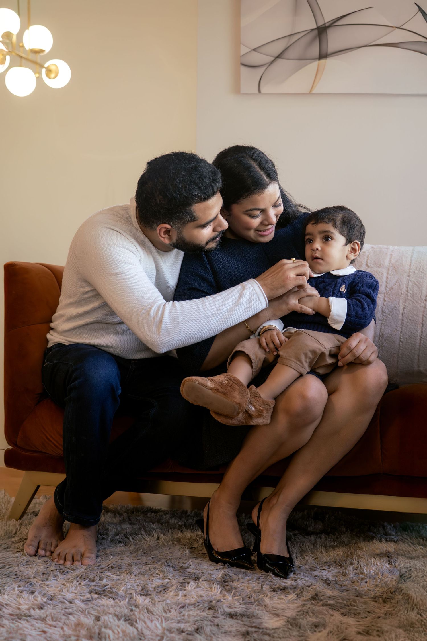 A happy family of three sits on a brown sofa, parents smiling at their toddler in a cozy living room.