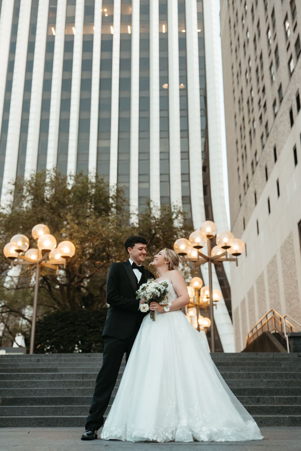 Bride and groom embrace on urban steps with city skyscrapers and glowing lamp posts behind them.