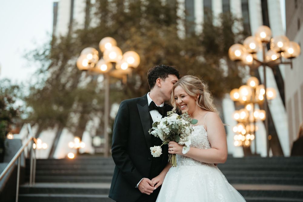 Bride and groom share romantic moment on steps, groom in tuxedo kissing brides cheek as she holds white floral bouquet.