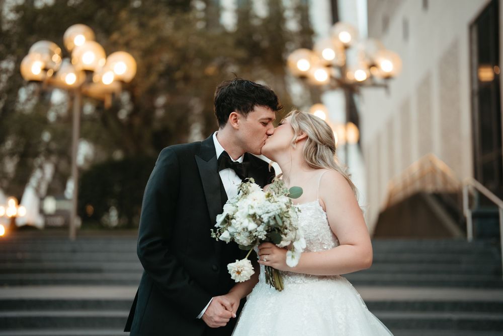 Bride and groom share a romantic kiss on steps, bride holding white bouquet, groom in black tuxedo, warm lights behind.