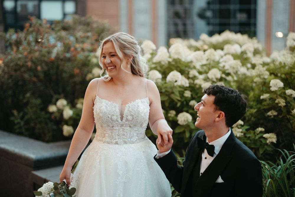 Bride in lace ballgown smiles at groom in black tuxedo outdoors with white hydrangeas in background.