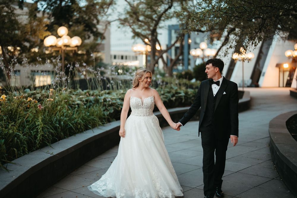 Bride in white ballgown and groom in black tuxedo hold hands walking through an elegant outdoor garden venue.