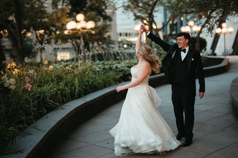 Bride and groom dancing outdoors at dusk, groom in tuxedo spinning bride in white ballgown near garden path.