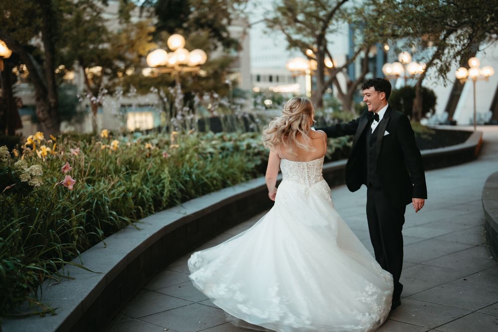 Bride in white ball gown and groom in black tux walk hand in hand through an elegant garden at dusk.