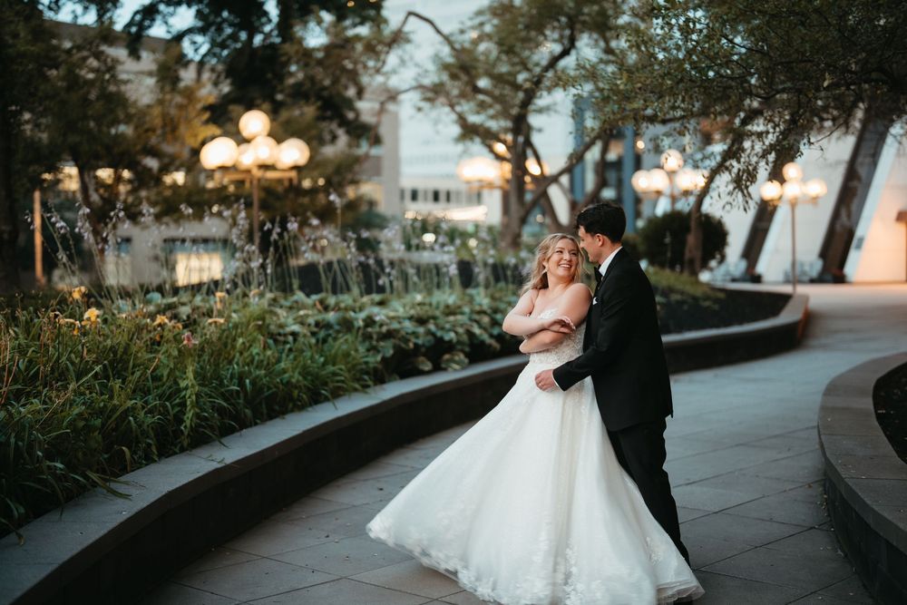 A bride and groom share a romantic embrace on a curved pathway surrounded by lush gardens and glowing lanterns at dusk.