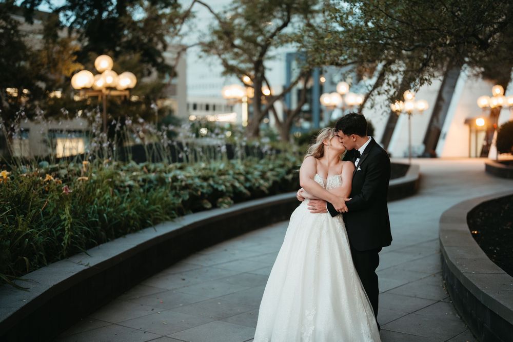 Bride and groom share a romantic kiss in an elegant outdoor garden setting at dusk, surrounded by glowing lights.