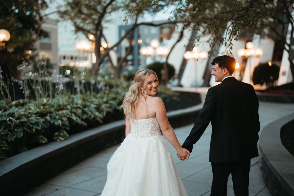 A bride and groom hold hands while walking through an elegant outdoor garden venue at dusk.