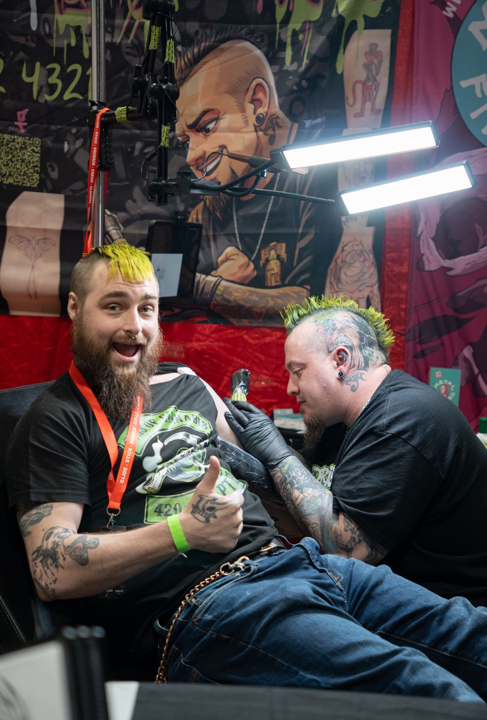 Two tattooed men with colorful mohawks at a tattoo convention, one getting a tattoo while smiling at the camera.