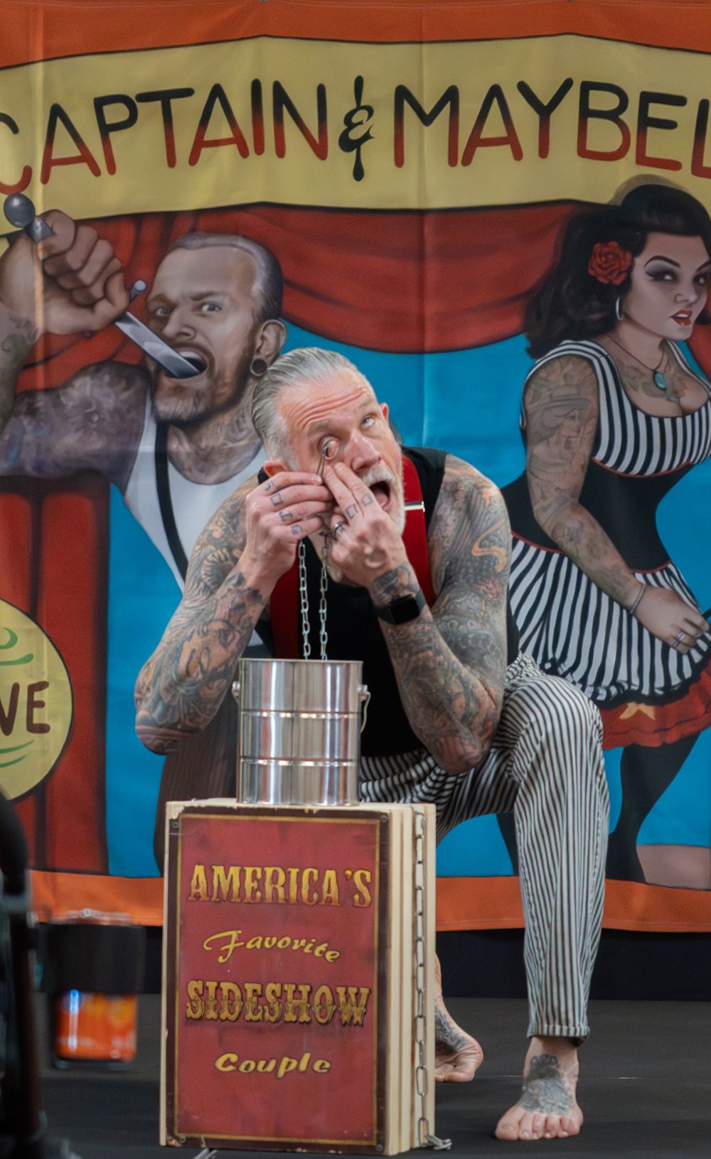Tattooed sideshow performer on stage at America's Favorite Sideshow Couple act with Captain & Maybelle banner.