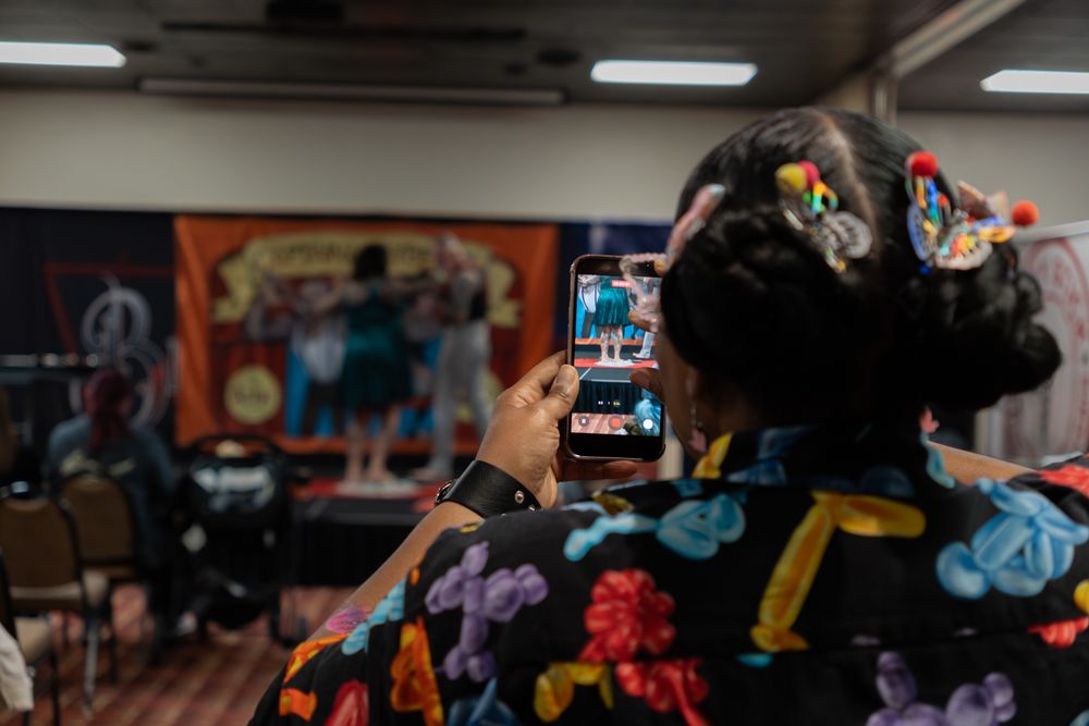 Woman in floral kimono with decorative hair clips photographs colorful comic art display with smartphone at convention.
