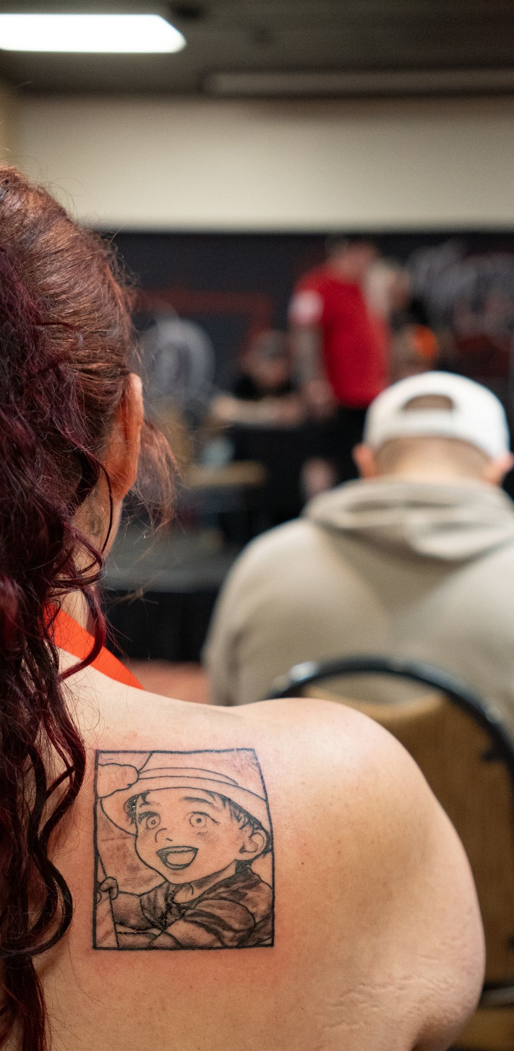 Woman with red hair showing a cartoon character tattoo on her upper back at an indoor event.