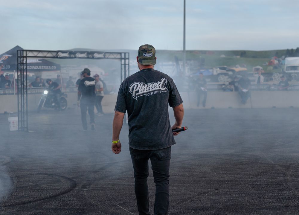 Man in Pinned t-shirt stands amid smoke at an outdoor motorsport burnout event with motorcycles in background.