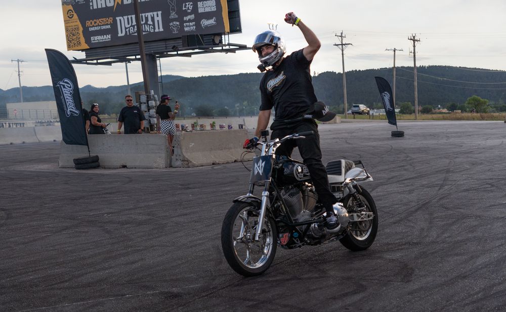 Motorcyclist performing a stunt on a custom chopper at an outdoor event, raising fist in triumph on a smoky asphalt track.