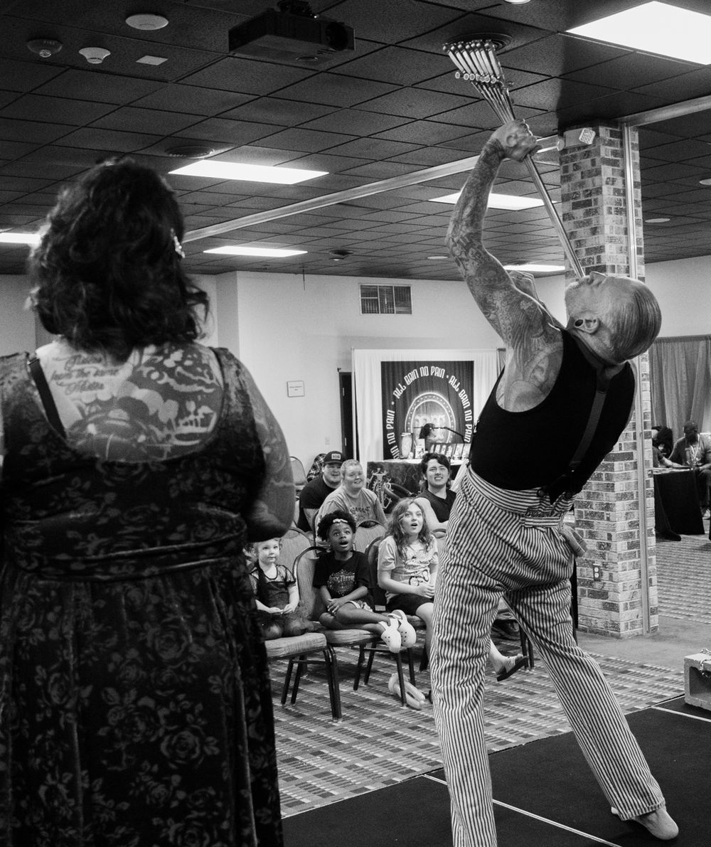 Tattooed performer raises a metal tool overhead before an audience in a black and white indoor show.
