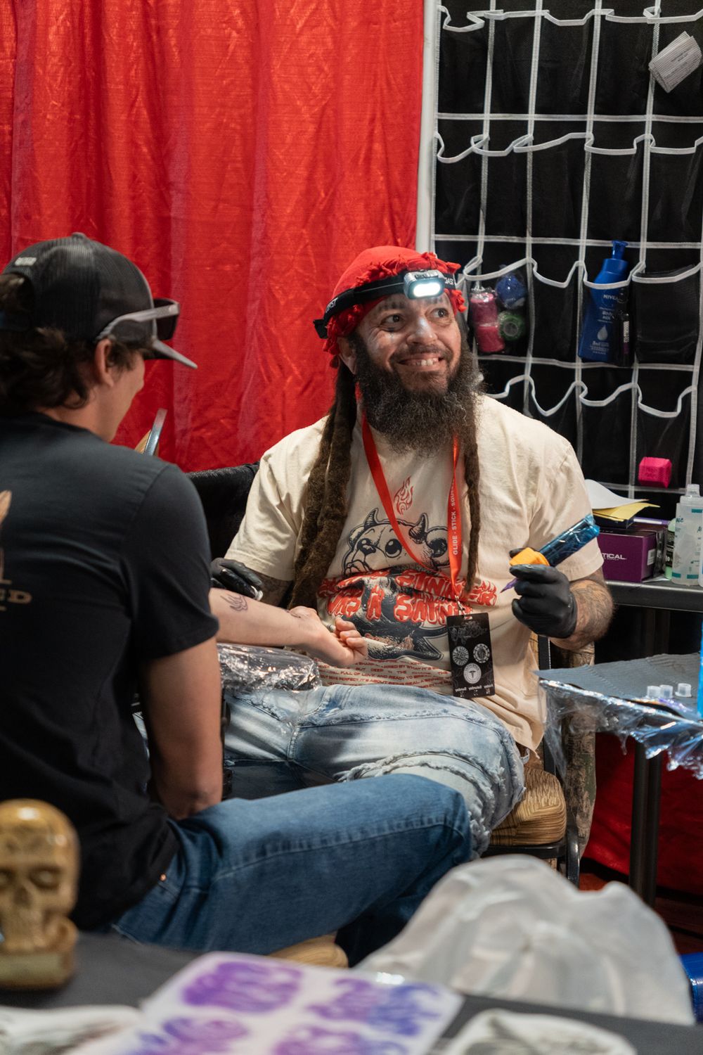 Tattoo artist with dreadlocks and red hat smiling while working at a tattoo convention booth.