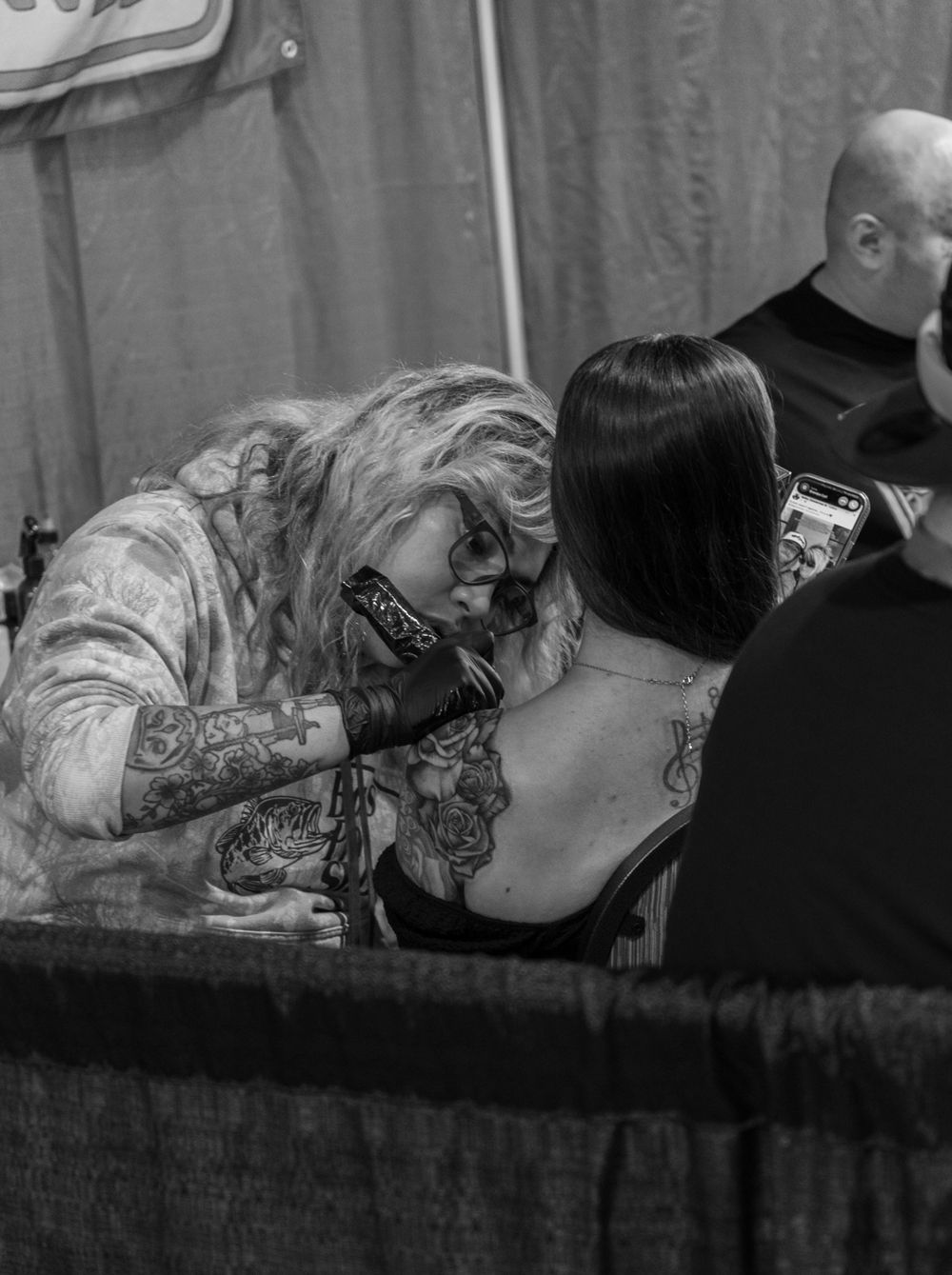 Black and white photo of a heavily tattooed artist giving a tattoo on a womans back at a tattoo convention.