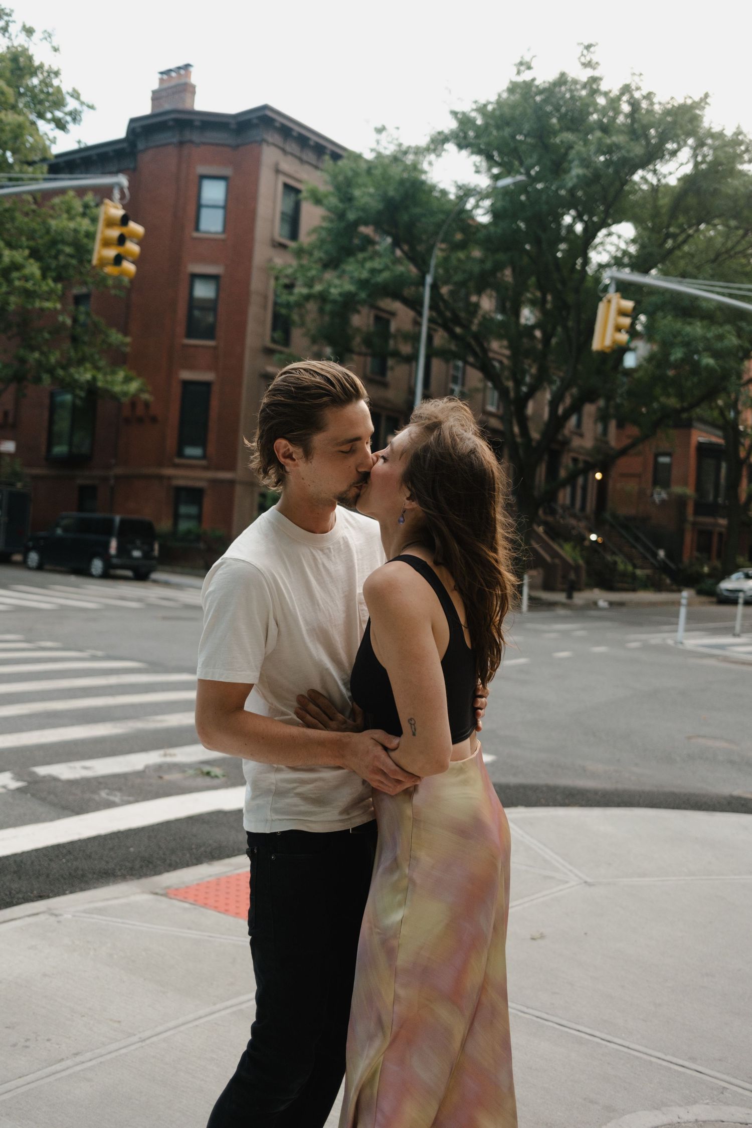 Couple sharing a romantic kiss on a NYC street corner, woman in floral skirt, man in white tee, brick buildings behind.