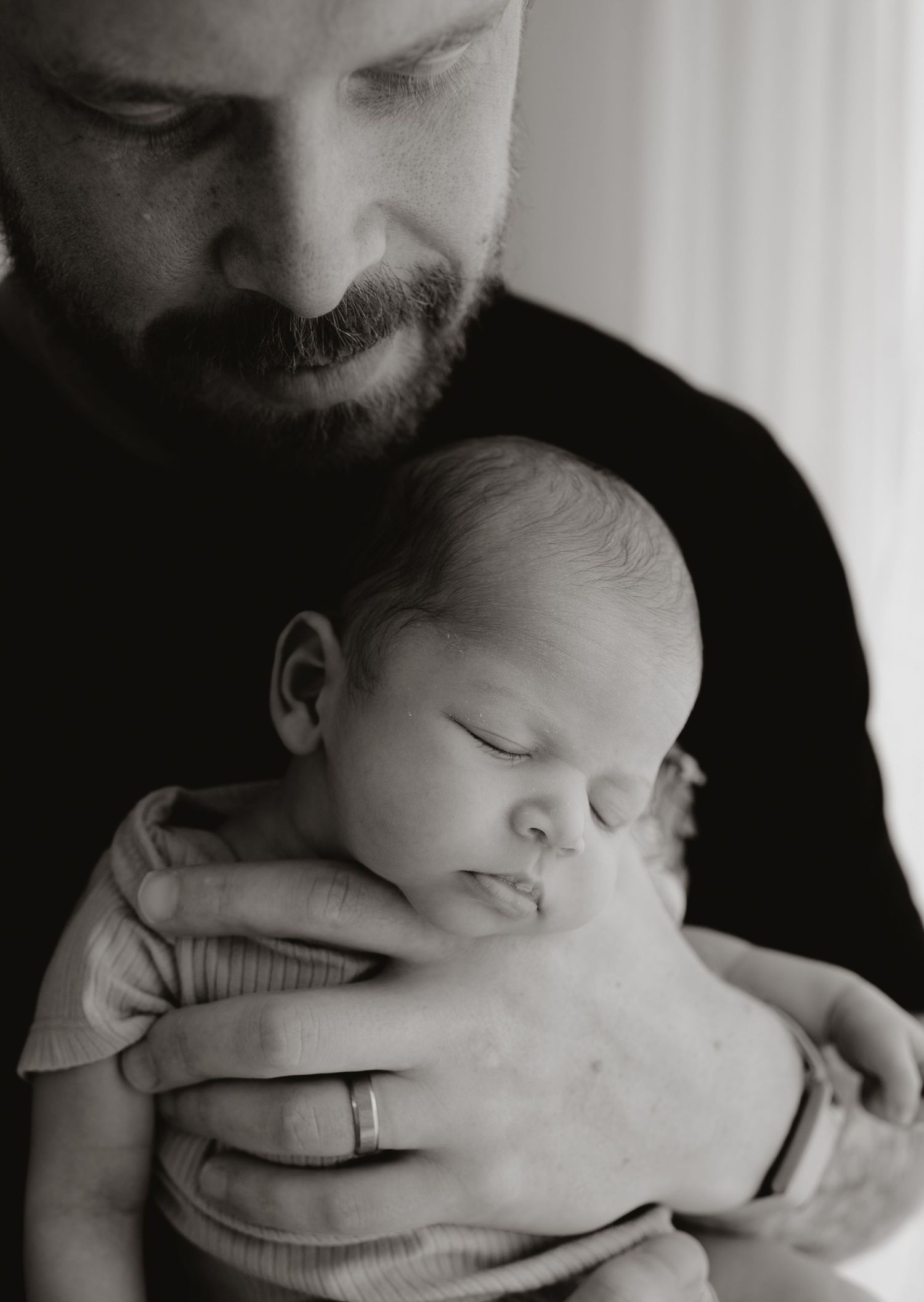 Black and white photo of a bearded father cradling his sleeping newborn baby, wearing a wedding ring.
