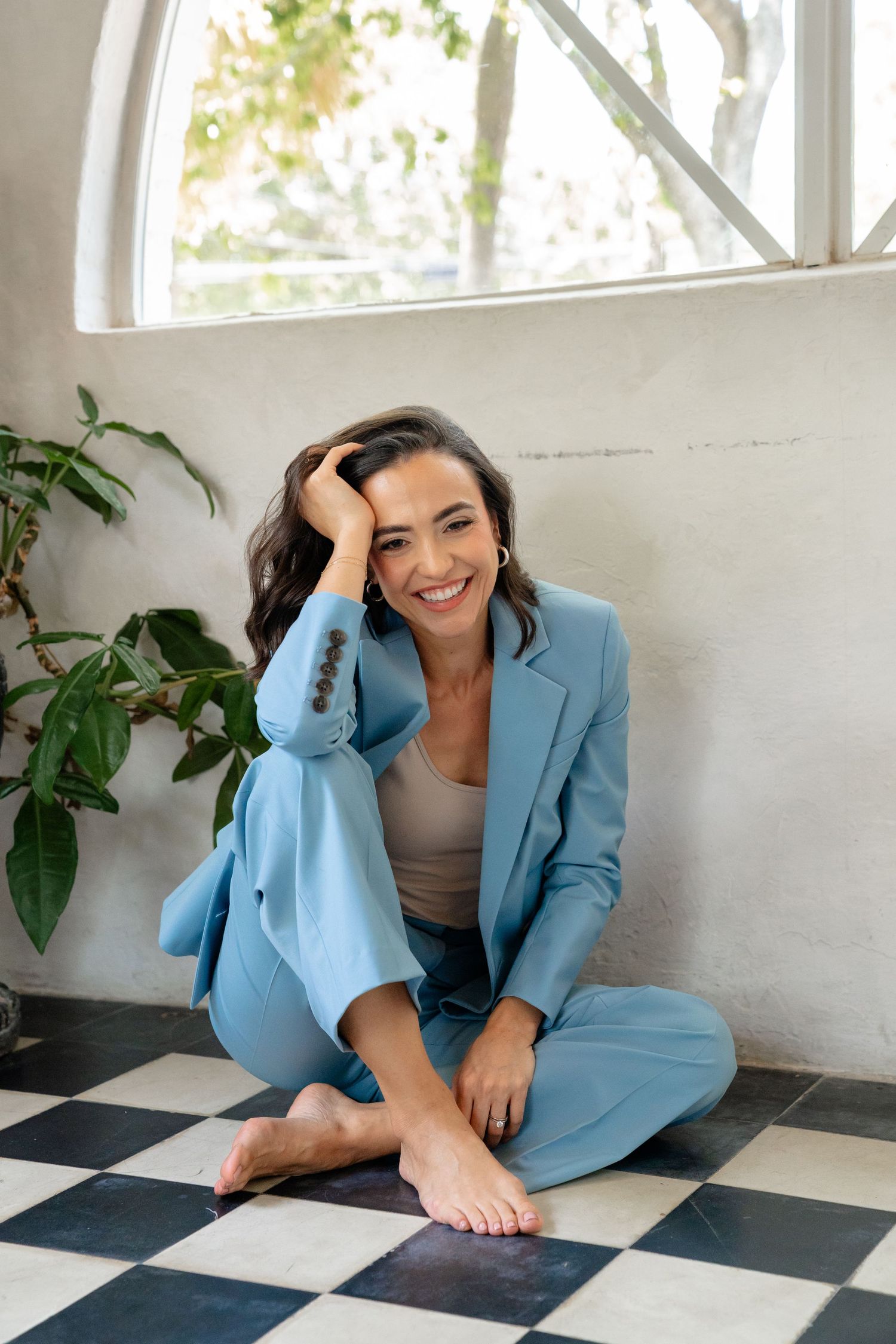 Smiling woman in blue suit sitting cross-legged on checkered floor near window and plant.