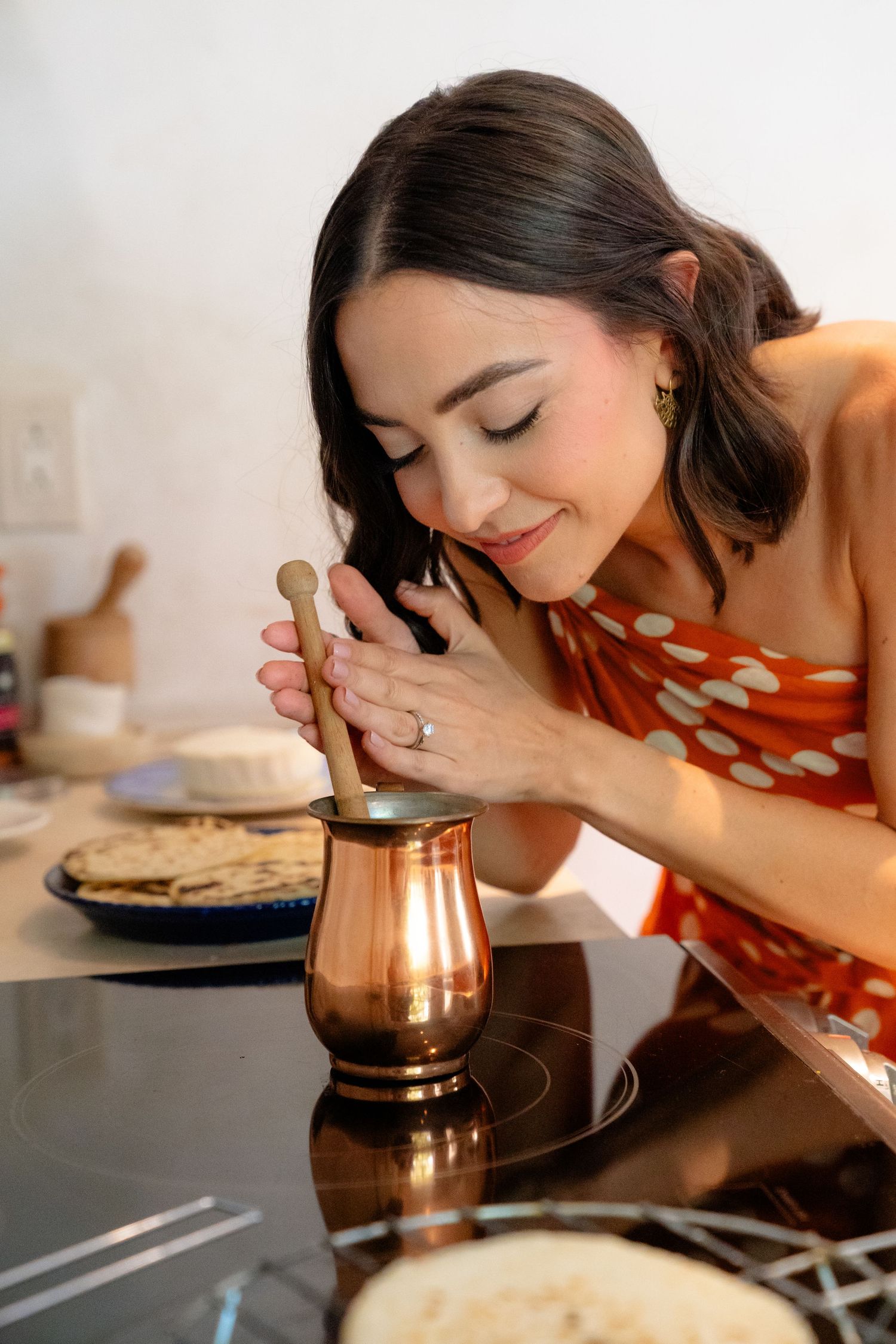 Woman in red floral dress smiling while using a copper mortar and pestle in a modern kitchen.