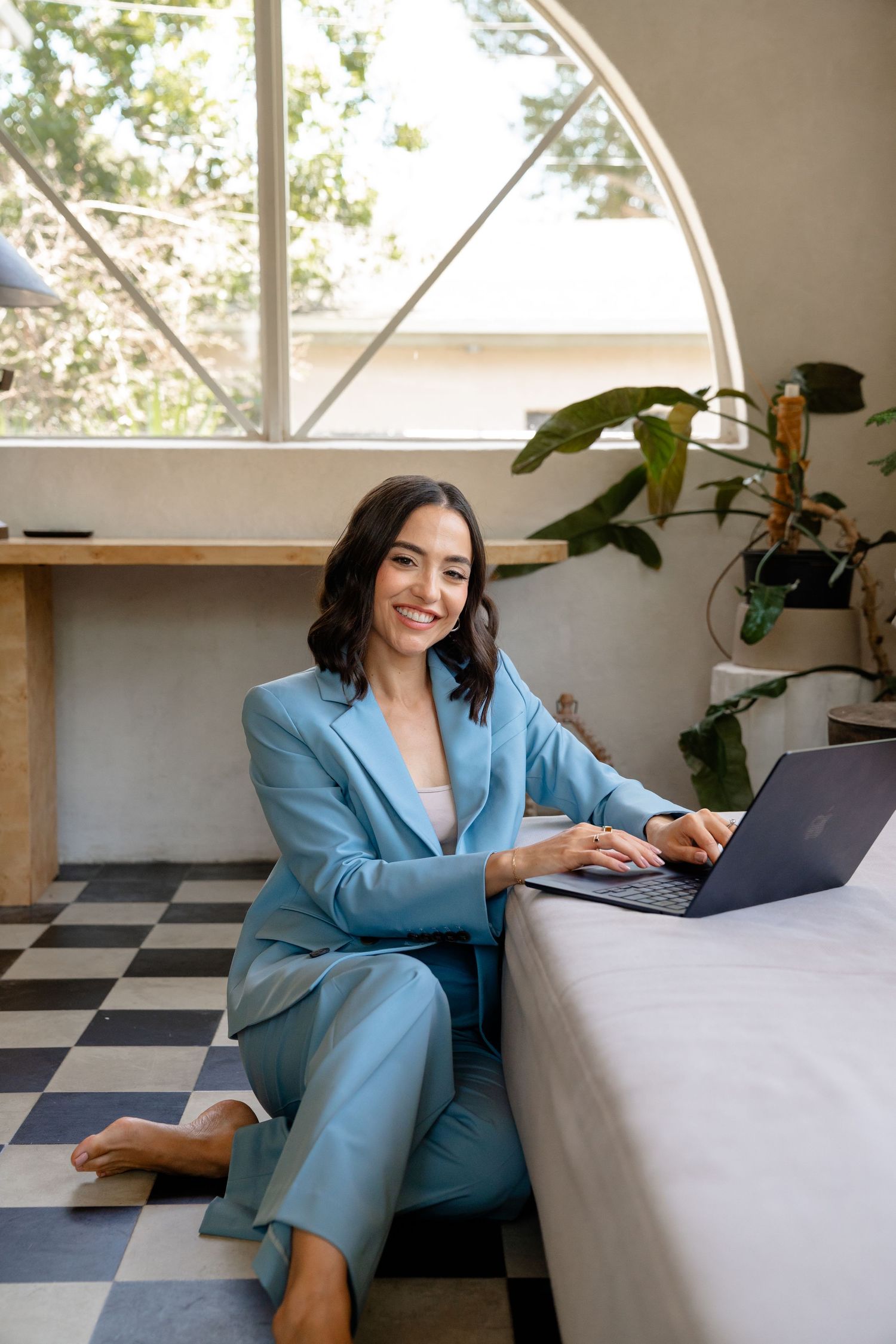 Woman in blue suit sitting on floor with laptop, smiling in modern room with checkered floor and plants.