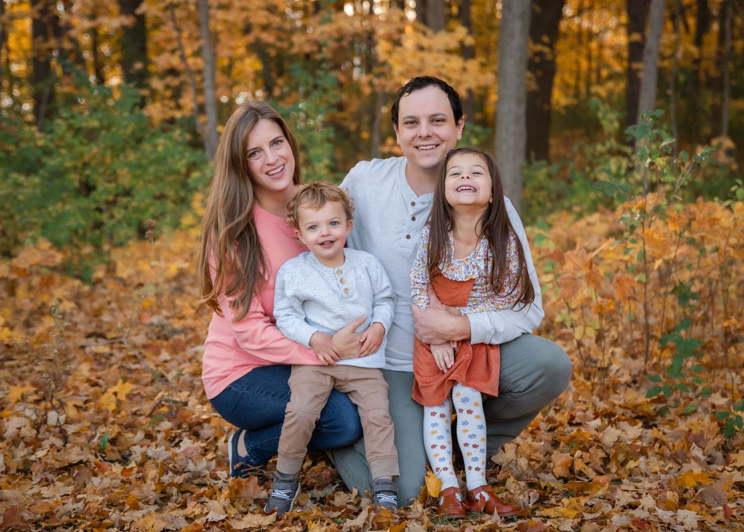 Family of four smiling in autumn forest with golden leaves, parents crouching with two young children in fall outdoor portrait.