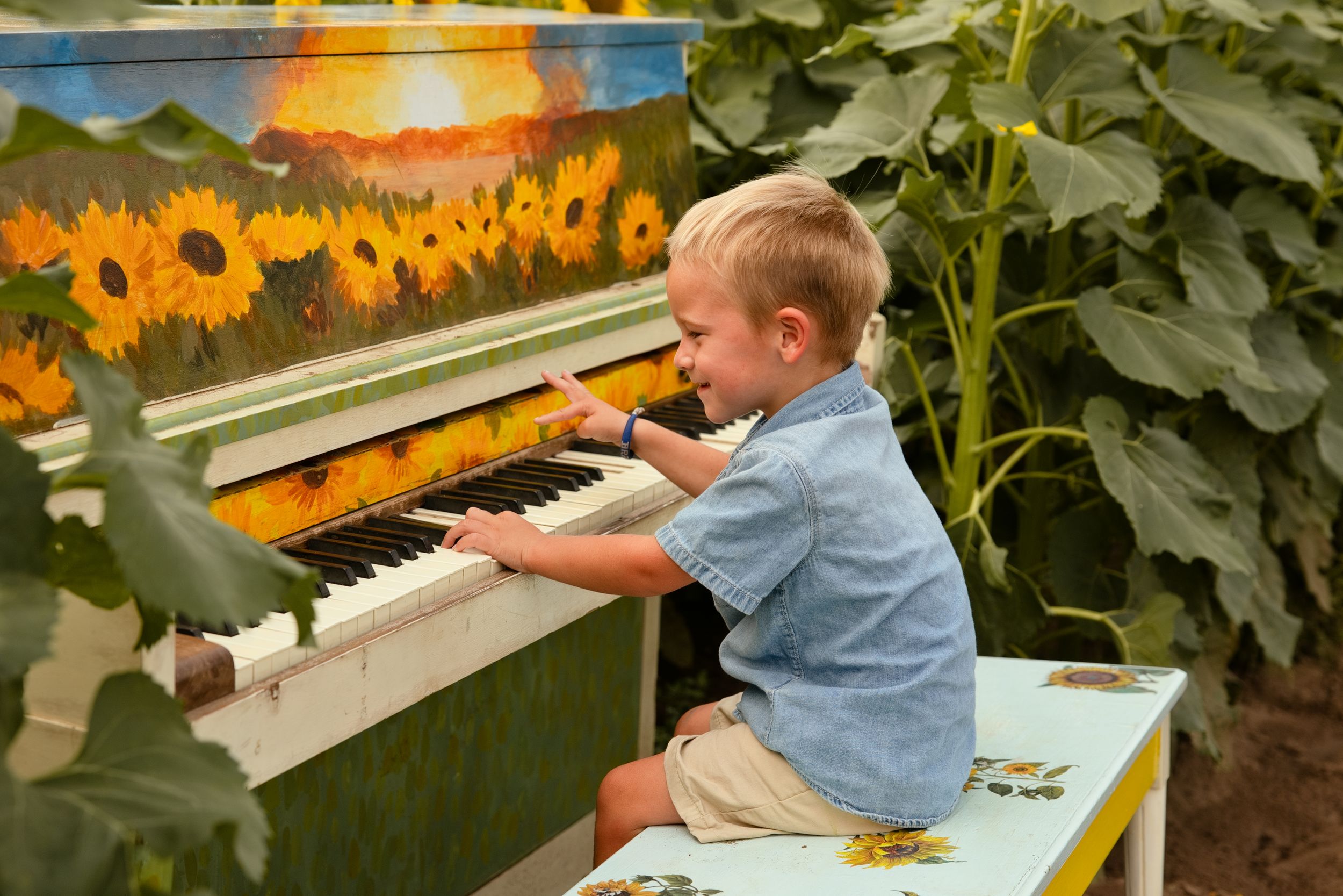 A joyful toddler boy plays a sunflower-painted upright piano outdoors surrounded by tall sunflowers.