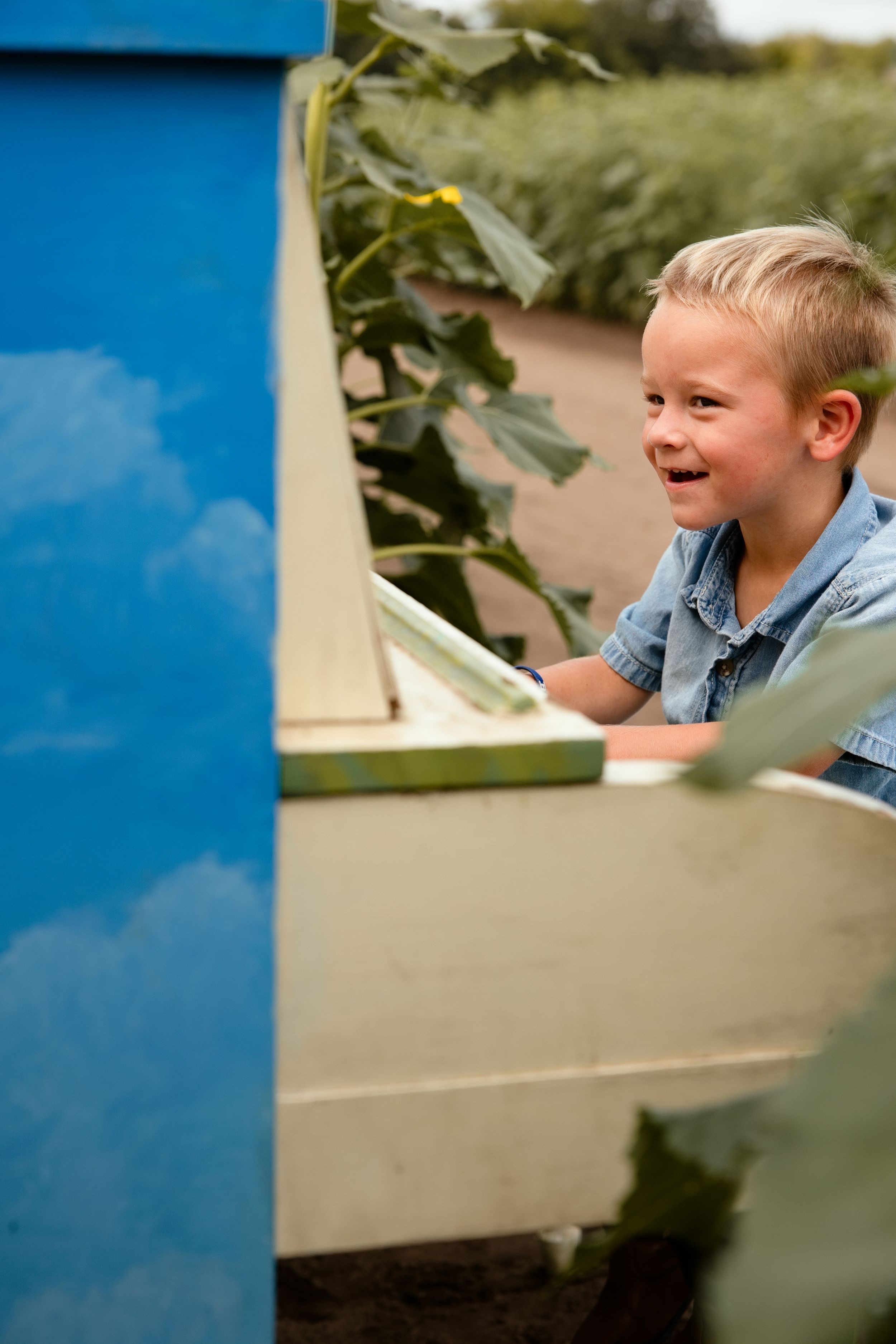 A happy young blonde boy in a denim shirt laughing outdoors near a blue and white wooden structure in a garden.