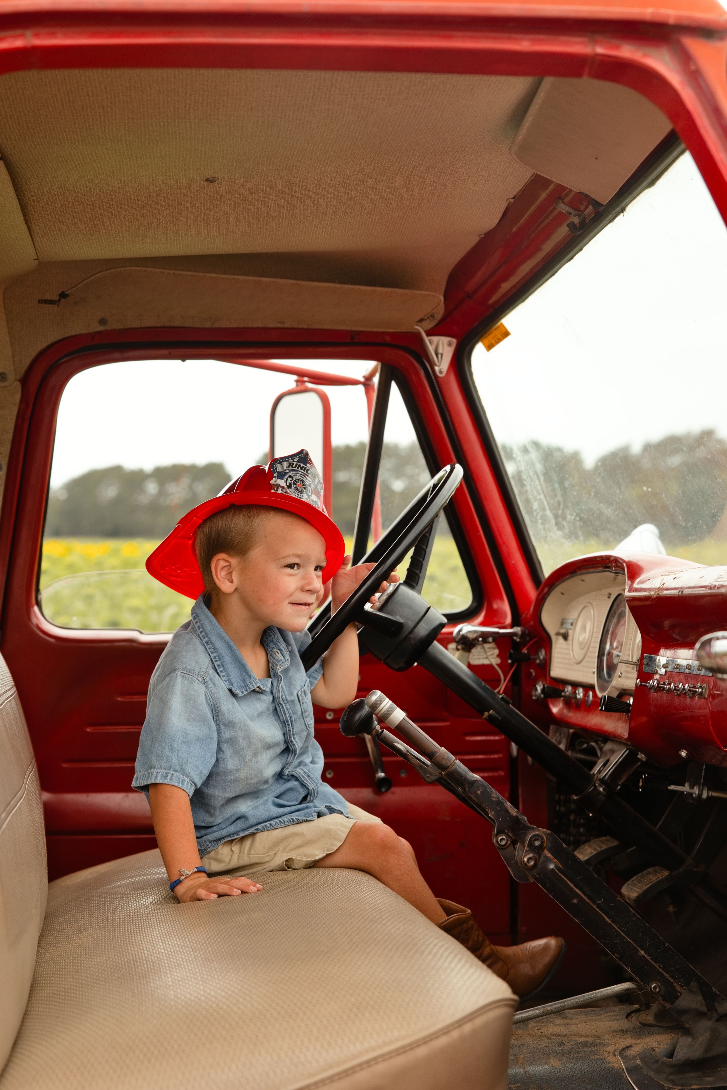 A smiling young boy wearing a red firefighter hat sits in the cab of a vintage red fire truck.