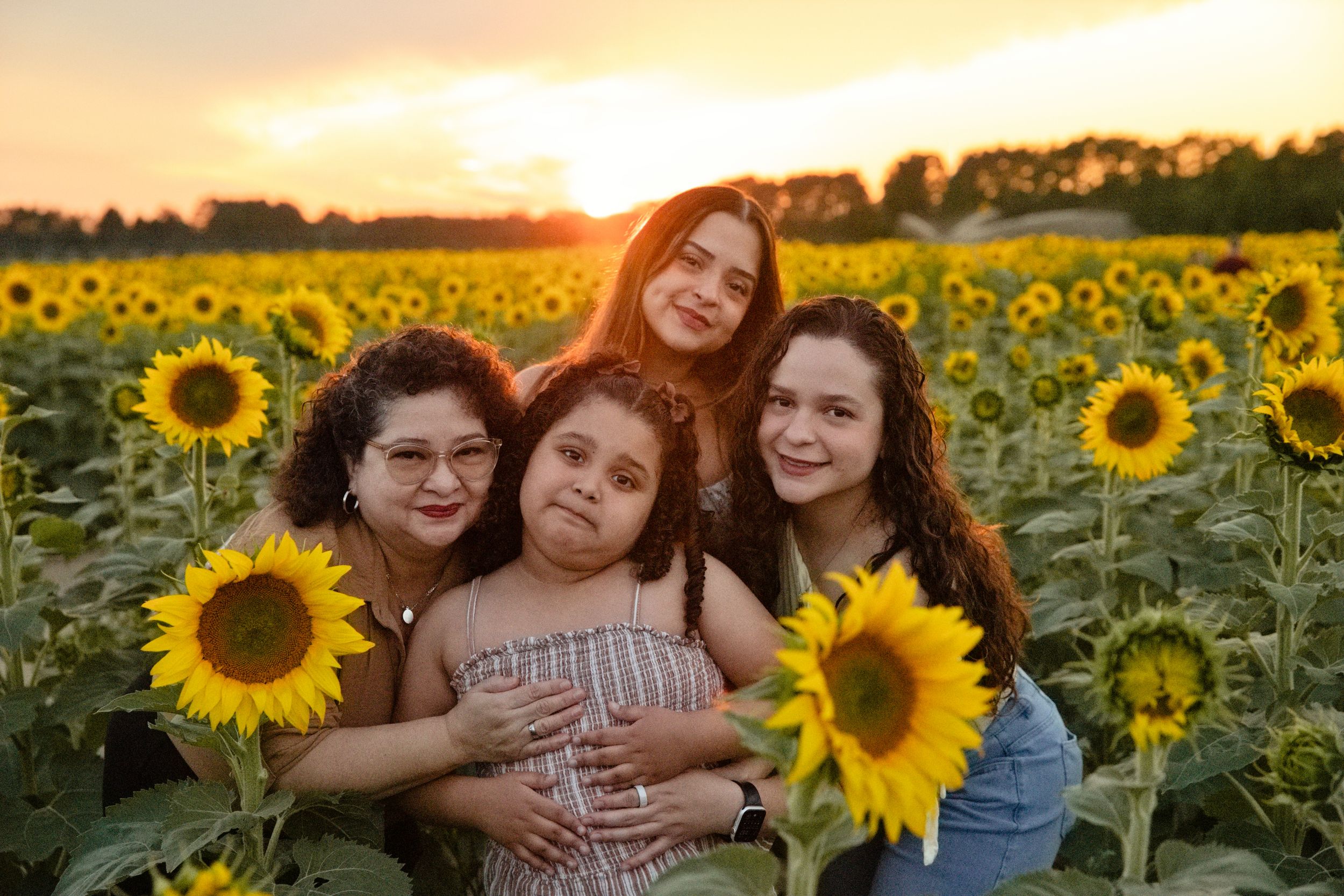Four girls pose together in a vibrant sunflower field at golden sunset, smiling warmly for a family portrait.