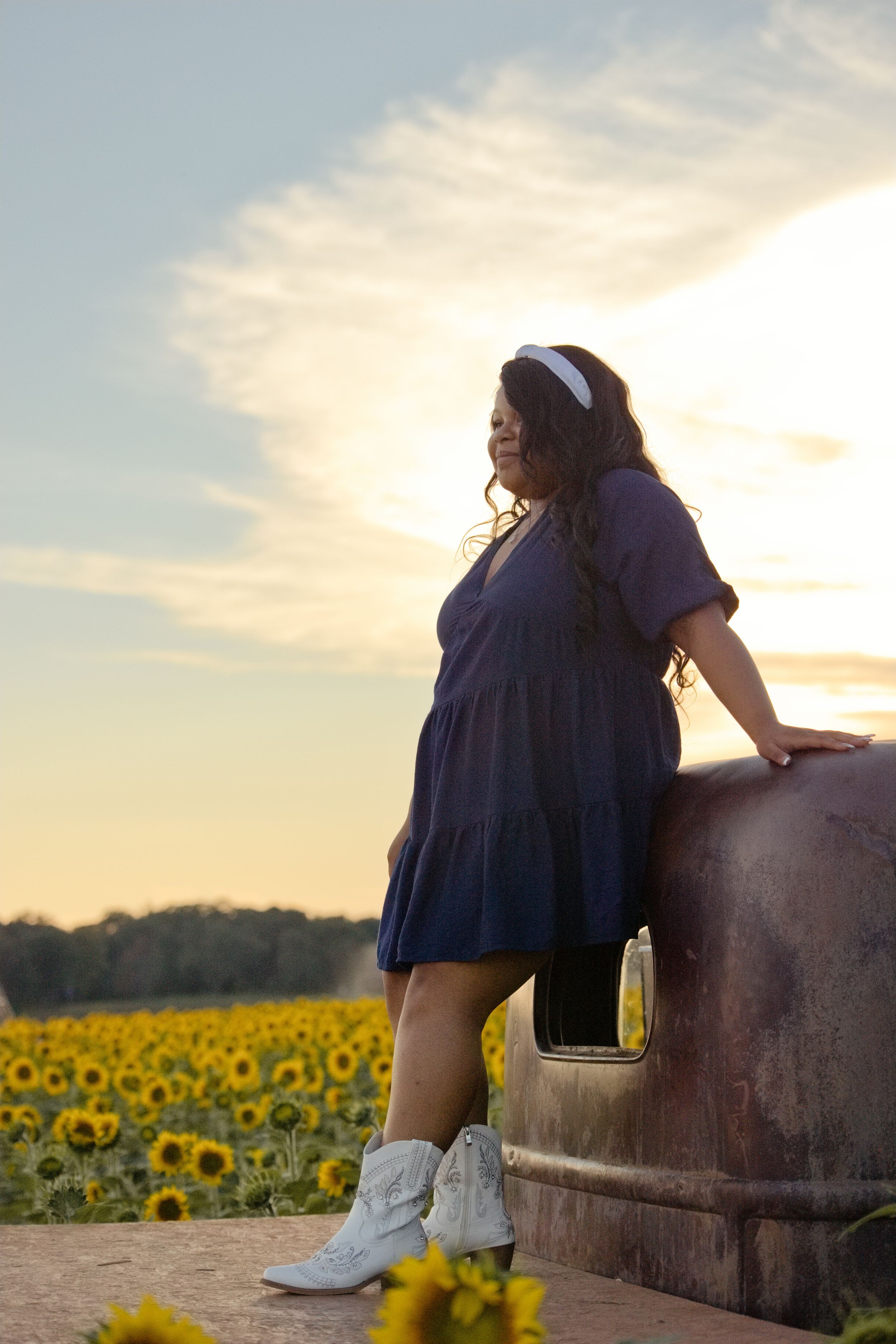 Woman in blue dress and white cowboy boots leaning on vintage truck in sunflower field at sunset.