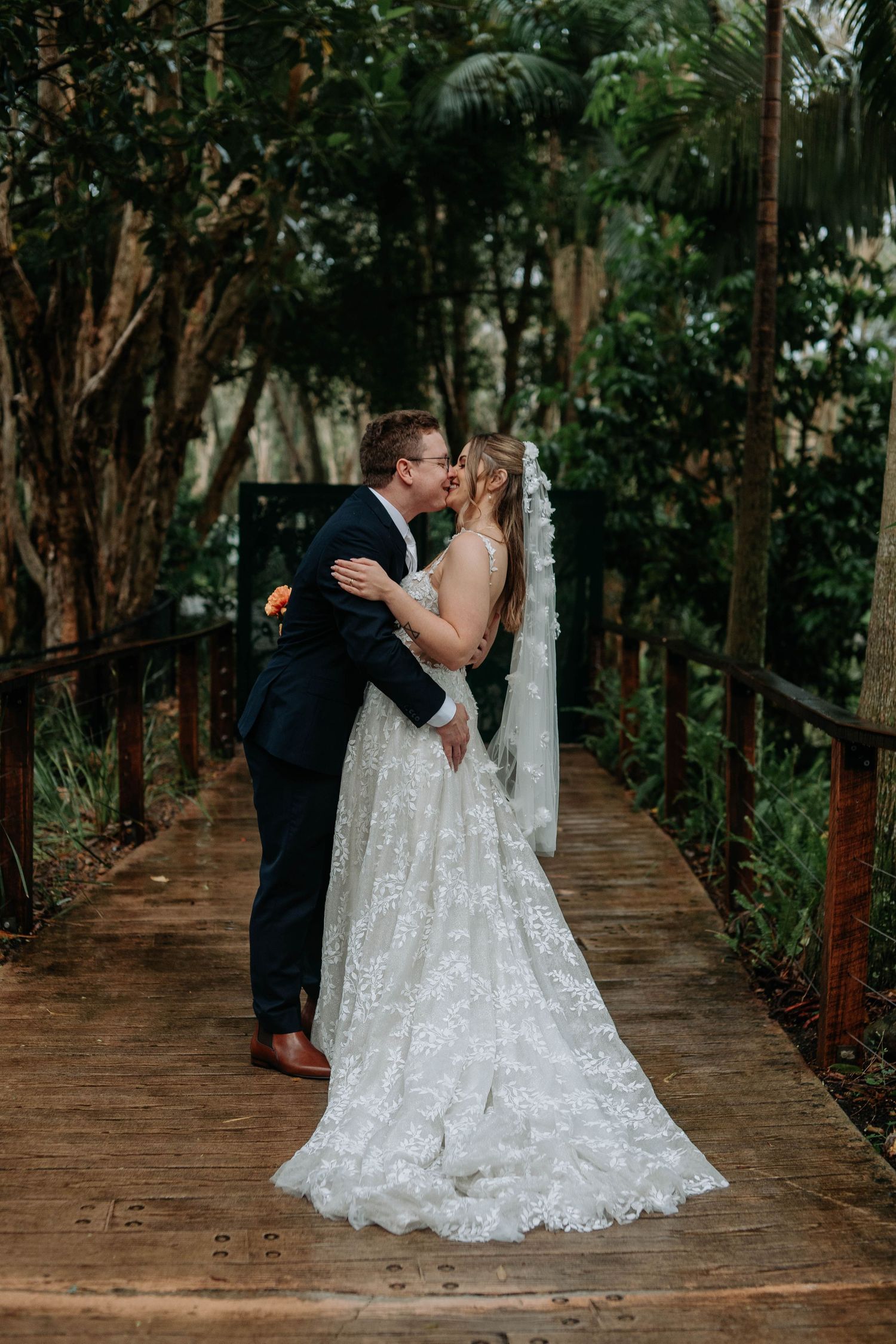 A bride and groom share a romantic kiss on a wooden bridge surrounded by lush tropical greenery on their wedding day.