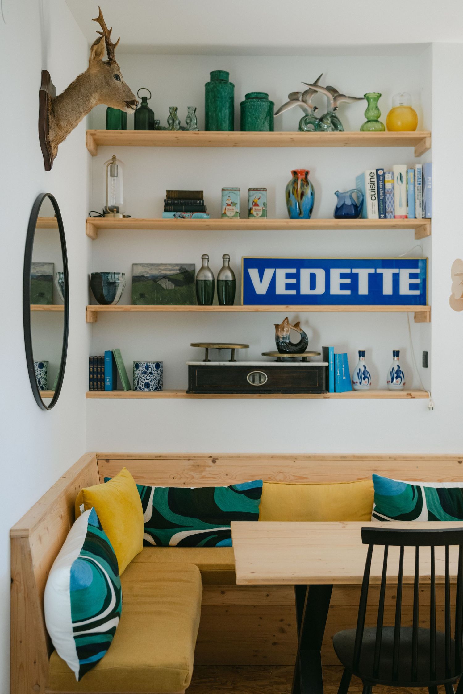 Modern dining room with long wooden table, colorful cushions, unique bubble chandelier, round mirror, and mounted deer head.