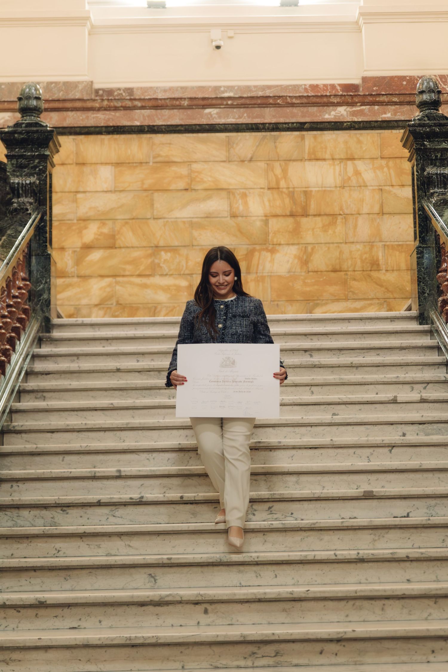 Young woman in tweed jacket walking down marble staircase while reading her diploma.