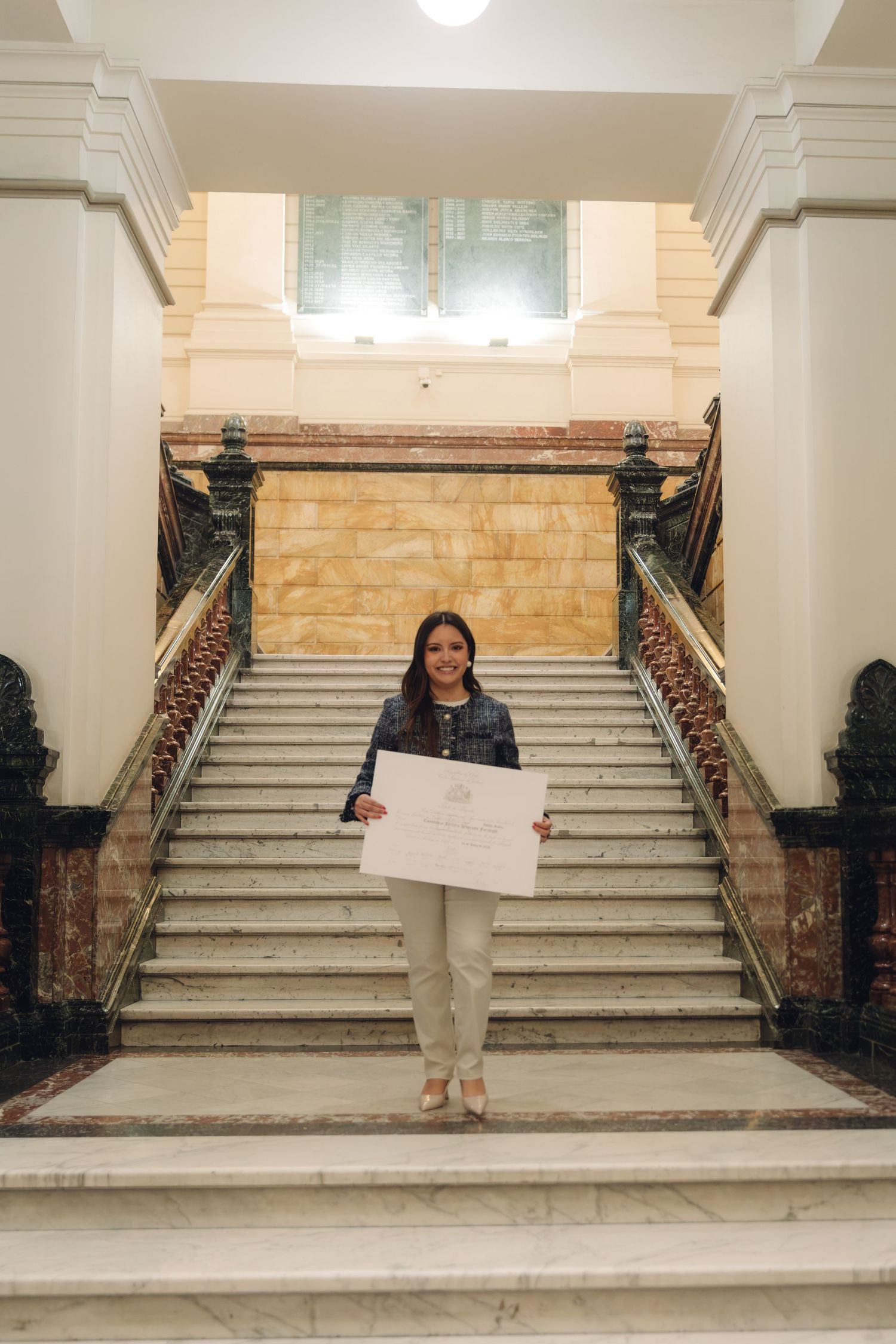 Smiling graduate displays diploma certificate on ornate marble staircase in classical building.