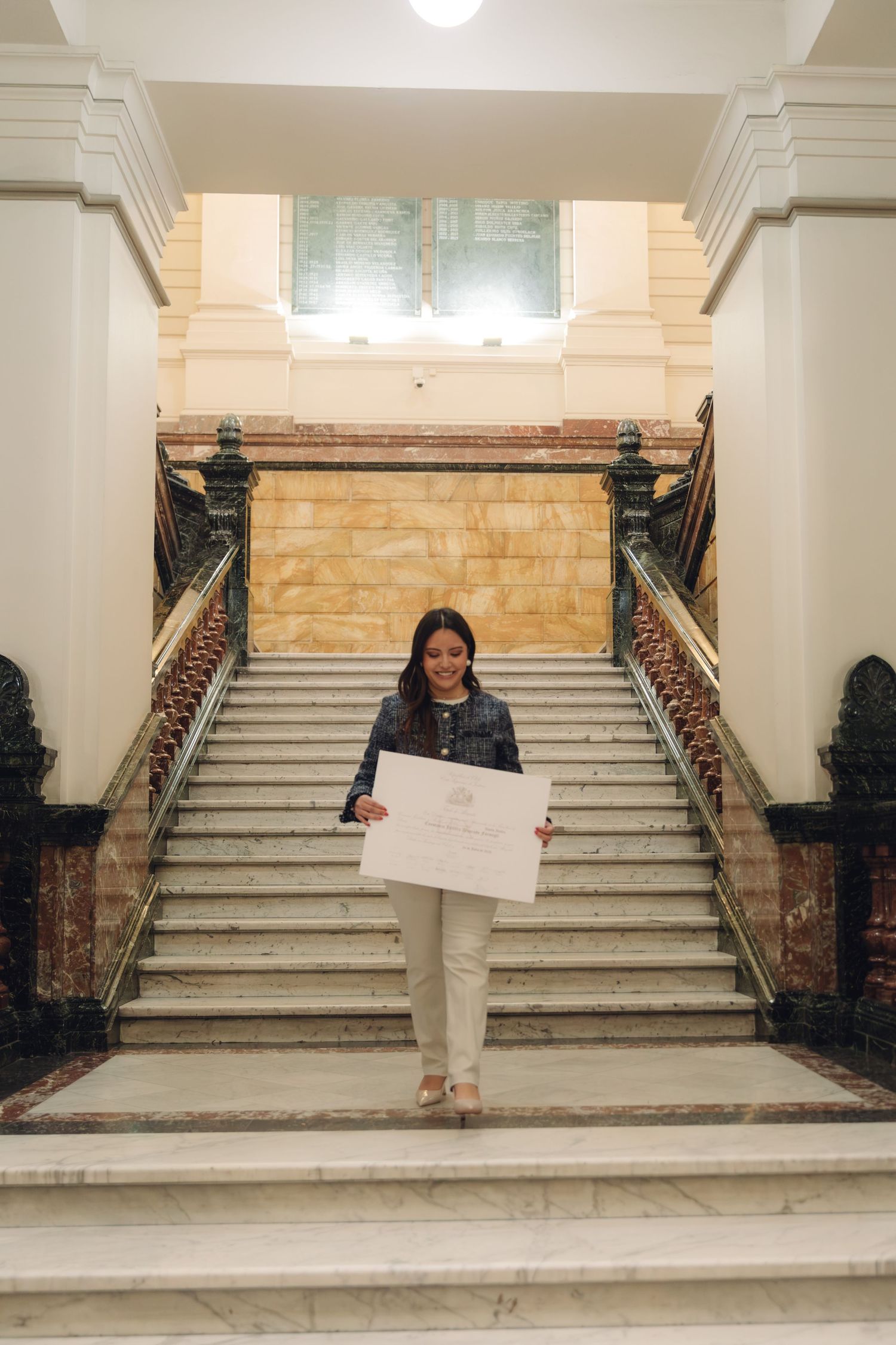 Young woman proudly holds diploma on grand marble staircase in elegant building.