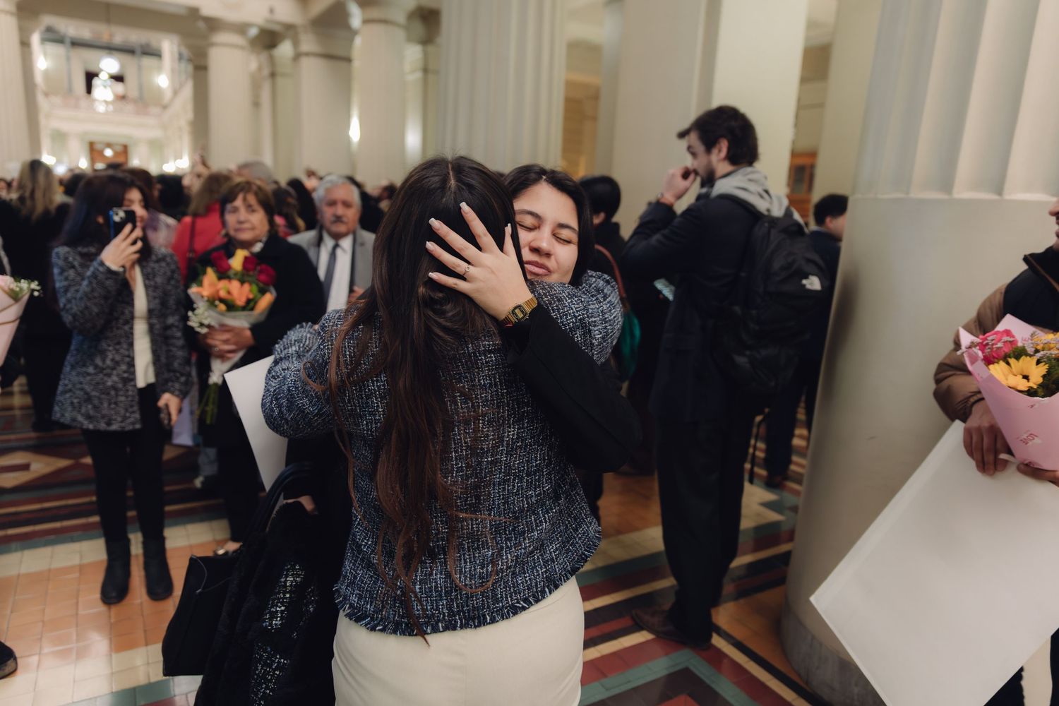 Two women share emotional hug at graduation ceremony in grand hall.