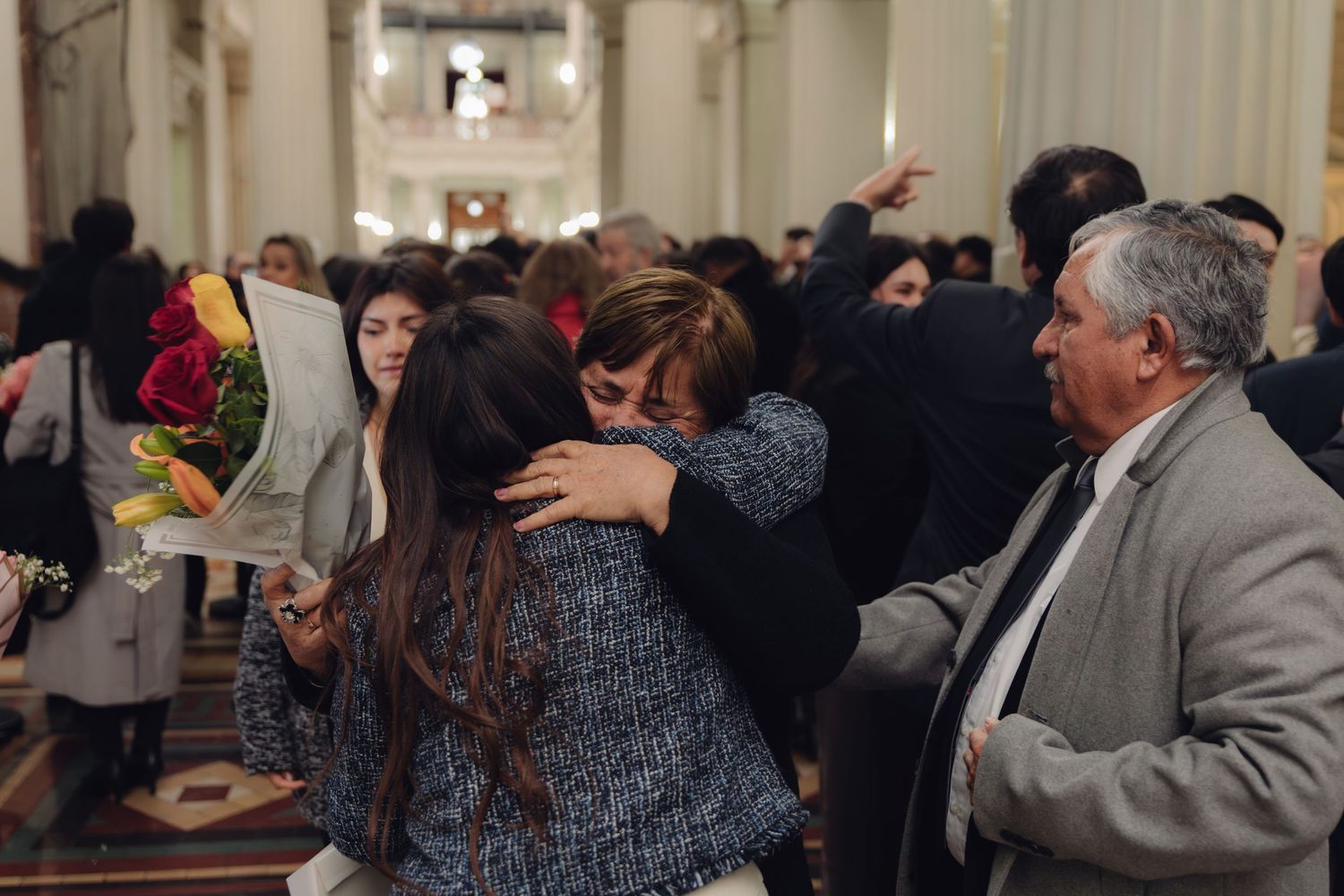 Family celebrates with hugs and flowers at university graduation ceremony indoors.