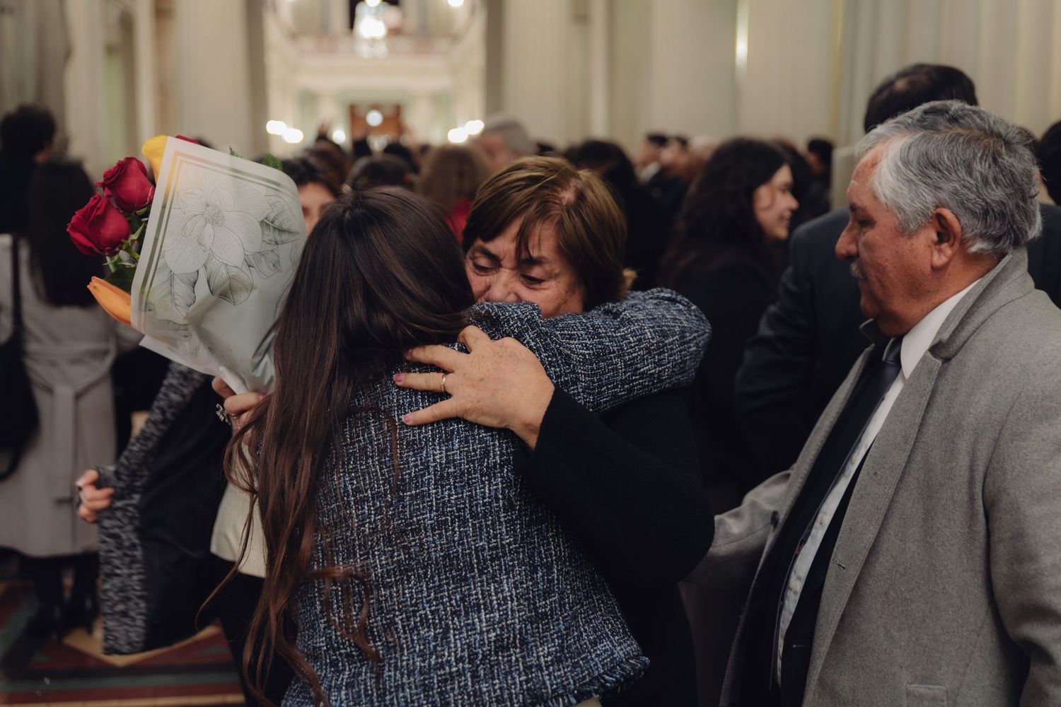 Graduate receives warm embrace from older woman while man watches at ceremony.