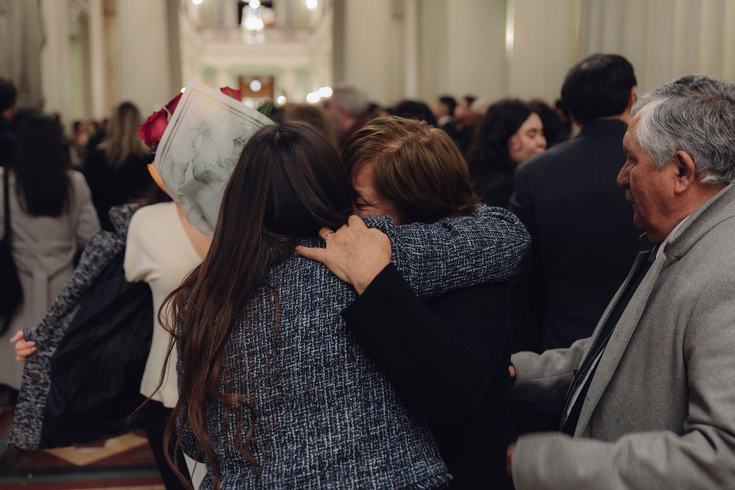 Graduate hugs family member in crowded ceremonial hall during graduation celebration.