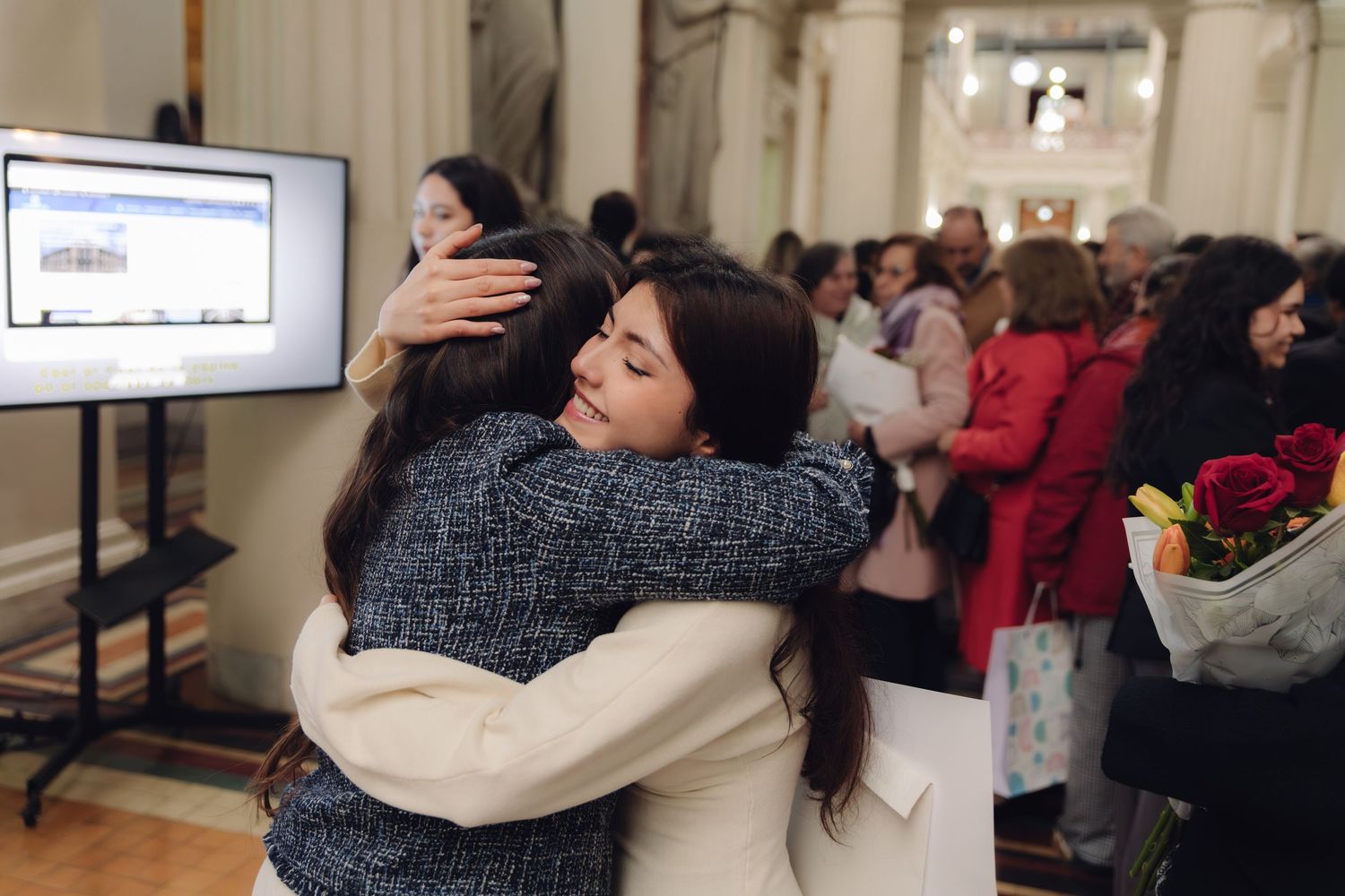 Two young women share heartfelt hug at graduation ceremony with diploma displayed nearby.